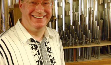 The photo shows Organist Dr Peter King sitting in front of a set of silver organ pipes. He is wearing a black and white shirt.