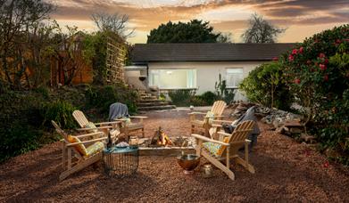 A cosy garden space at sunset featuring chairs arranged around a warm fire pit, perfect for gatherings and relaxation.. There is a white house in the